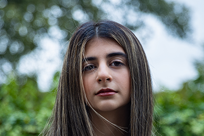 A young woman with dark brown hair and blonde highlights is looking into the camera with a powerful gaze.