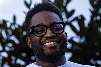 Rhammel is a young black man with short cropped hair wearing a fitting blue shirt and big black glasses. He is standing in front of fields soaked in sunlight.