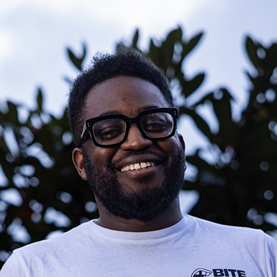 Rhammel is a young black man with short cropped hair wearing a fitting blue shirt and big black glasses. He is standing in front of fields soaked in sunlight.
