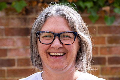 Ruth a middle aged white woman with shoulder length grey hair streaked with brown and big square dark blue glasses and wearing a blue and white striped shirt is standing in front of a blurry green and brown background.