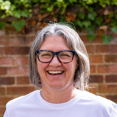 Ruth a middle aged white woman with shoulder length grey hair streaked with brown and big square dark blue glasses and wearing a blue and white striped shirt is standing in front of a blurry green and brown background.
