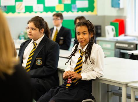 A young student wearing school uniform leans against a desk.