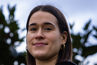 Sara has lightly tanned skin and brown hair pulled back into a sleek ponytail. She is wearing small stud earrings and smiling into the camera. She has a purple hoodie on with a black jacket on top.