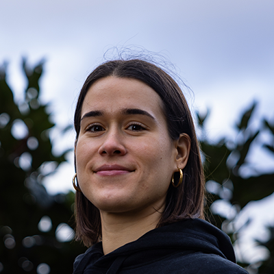 Sara has lightly tanned skin and brown hair pulled back into a sleek ponytail. She is wearing small stud earrings and smiling into the camera. She has a purple hoodie on with a black jacket on top.