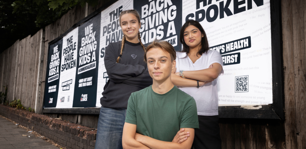 Three Bite Back activists Alice, Luke and Carrera are stood smiling in front of our Commercial Break Campaign posters.