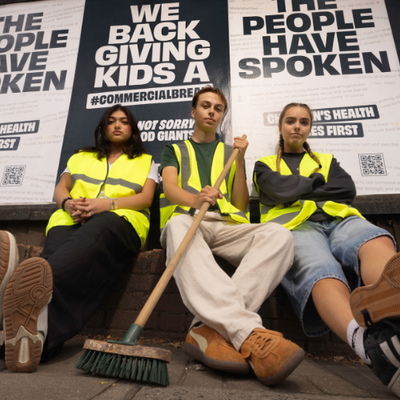 Three Bite Back activists sit infront of an outdoor billboard wearing high vis. They are looking down at the camera with defiant expressions. Their billboards behind them read The People Have Spoken We Back Giving Kids A Commercial Break