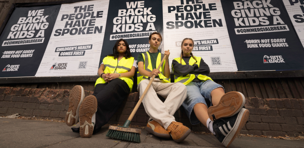 Three Bite Back activists sit infront of an outdoor billboard wearing high vis. They are looking down at the camera with defiant expressions. Their billboards behind them read The People Have Spoken We Back Giving Kids A Commercial Break