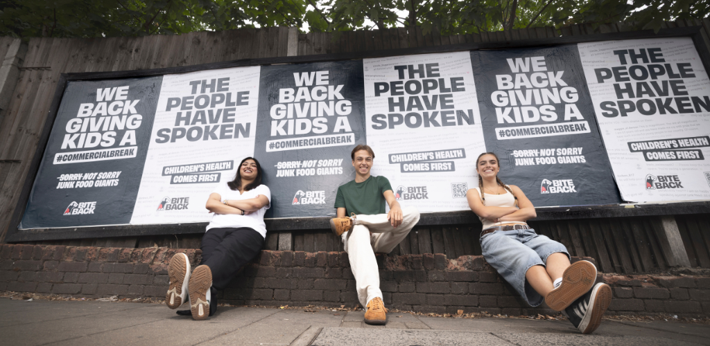Three Bite Back activists sit on a wall on a street in front of billboard posters, they have big smiles on their faces. The posters behind them read the people have spoken we back giving kids a commercial break