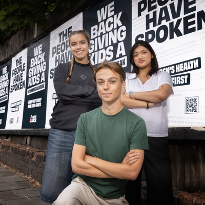 Three Bite Back activists stand on the street in front of billboard posters, they are smiling with their arms crossed. The posters behind them read the people have spoken we back giving kids a commercial break