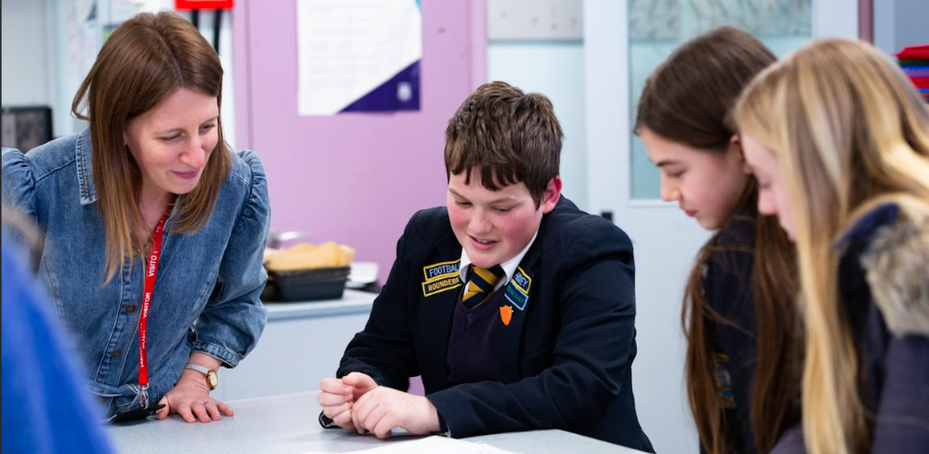 Three students sit around a classroom table with an adult, they are smiling and taking part in an activity