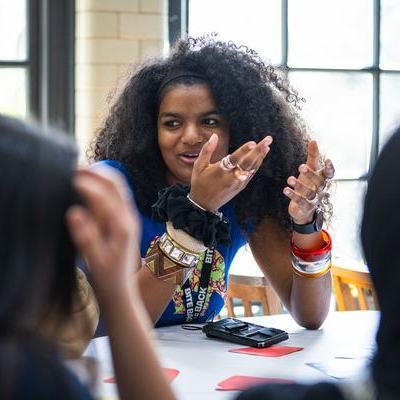 A young woman with curly hair animatedly speaks and gestures at a table during a lively group discussion. She wears colorful bracelets and a lanyard; cards and a phone lie on the table by a sunlit window.