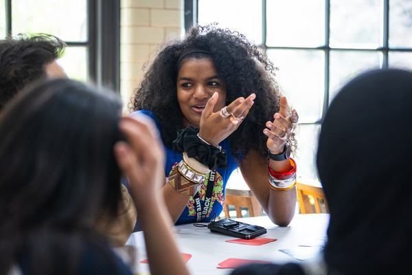 A young woman with curly hair animatedly speaks and gestures at a table during a lively group discussion. She wears colorful bracelets and a lanyard; cards and a phone lie on the table by a sunlit window.