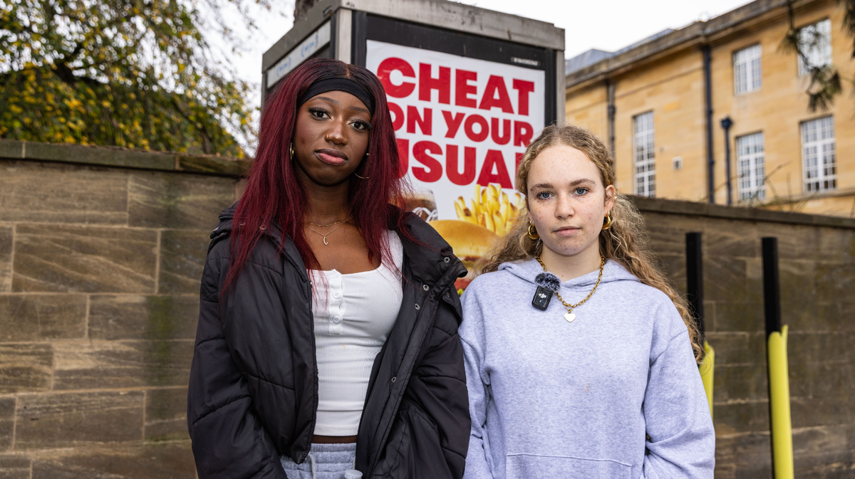 Two young people stand infront of a telephone box in Oxford with serious expressions. The telephone box has a huge poster advertising junk food, it reads cheat on your usual