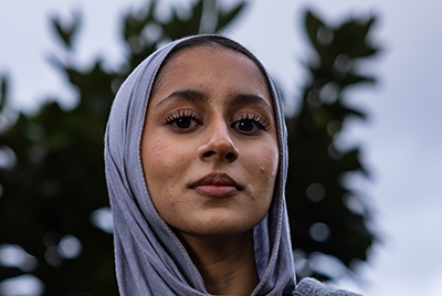 A young South Asian woman with a grey headscarf is looking down into the camera with a serious expression.