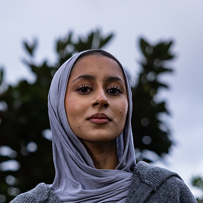 A young South Asian woman with a grey headscarf is looking down into the camera with a serious expression.