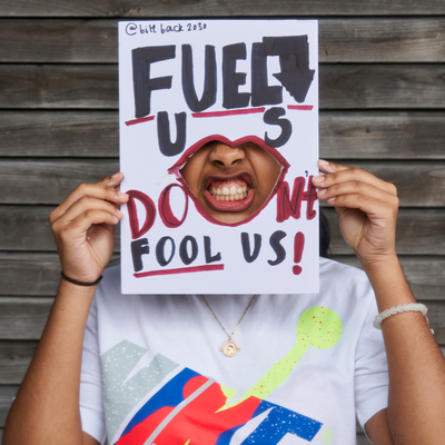 A brown skinned activist holding up a sign saying 'Fuel Us, Don't Fool Us' with a cut out through which she shows her mouth with bared teeth.