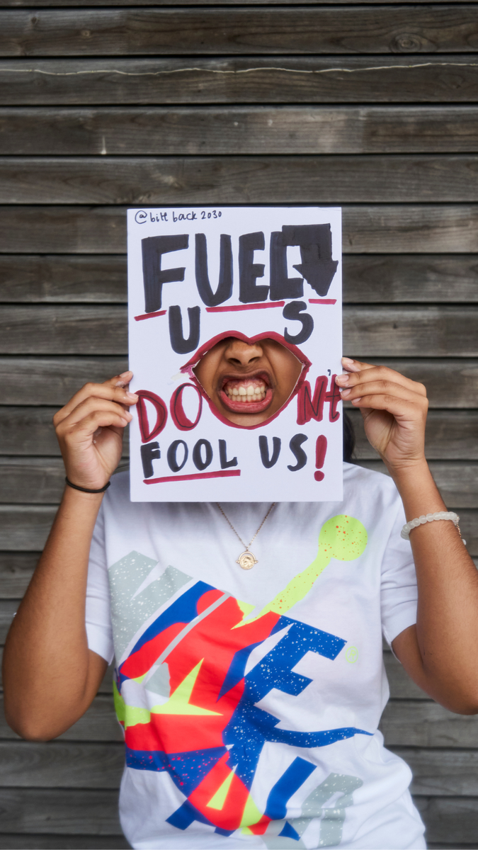 A brown skinned activist holding up a sign saying 'Fuel Us, Don't Fool Us' with a cut out through which she shows her mouth with bared teeth.