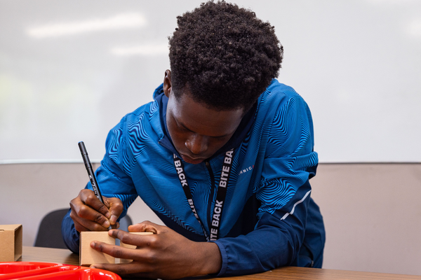 A young black man wearing a blue jacket is writing with a marker pen on a box