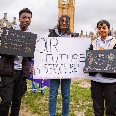 Timi standing together with two fellow activists holding a protest sign in front of Big Ben. He is a black young man with short cropped hair, wearing a black jacker and gray T-shirt holding a sign that reads "I believe in...putting Child Health First."