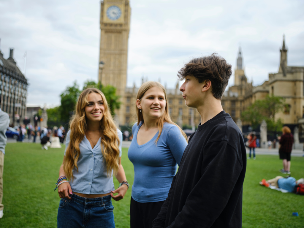 Three activists Alice, Maya and Oli P (left to right) standing on the parliament square in front of Westminster. Alice is a young white girl with long straight medium blonde hair, Maya is a white woman with shoulder length fair hair and Oli has messy brown hair that falls to his ears.