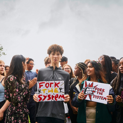A collection of youth board members holding protest signs and having fun laughing with each other. At the front is Vin, a pale young man with kinky brown longish hair. He's dressed in a grey Adidas sweatshirt and black slacks and holds a sign saying 'Fork The System' in red and yellow writing.
