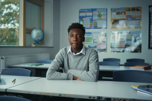 Young activist Timi, a slender young black man with shortish hair, is sitting at a table in a classroom. He is wearing a white suit shirt under a grey woolen jumper. Behind him are school posters from projects and there is a window on the left hand side.