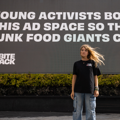YA young activist stands in front of a large digital billboard reading, “Young activists bought this ad space so that junk food giants couldn’t,” with the Bite Back logo visible.