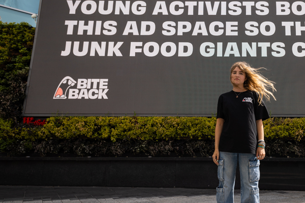 YA young activist stands in front of a large digital billboard reading, “Young activists bought this ad space so that junk food giants couldn’t,” with the Bite Back logo visible.