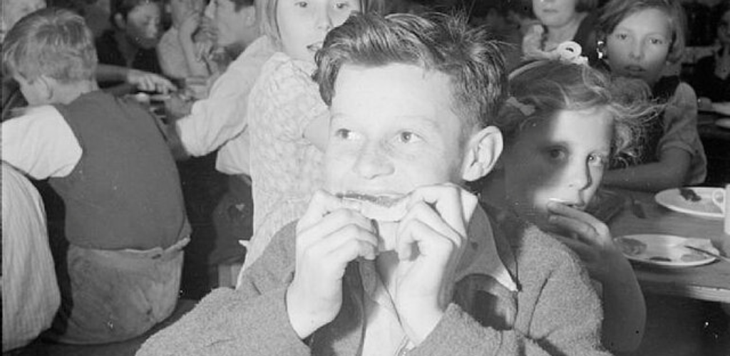 Black-and-white photo of children eating in a crowded dining hall. A smiling boy in the foreground bites into a sandwich while a girl behind him looks toward the camera; plates and cups are on the tables around them.