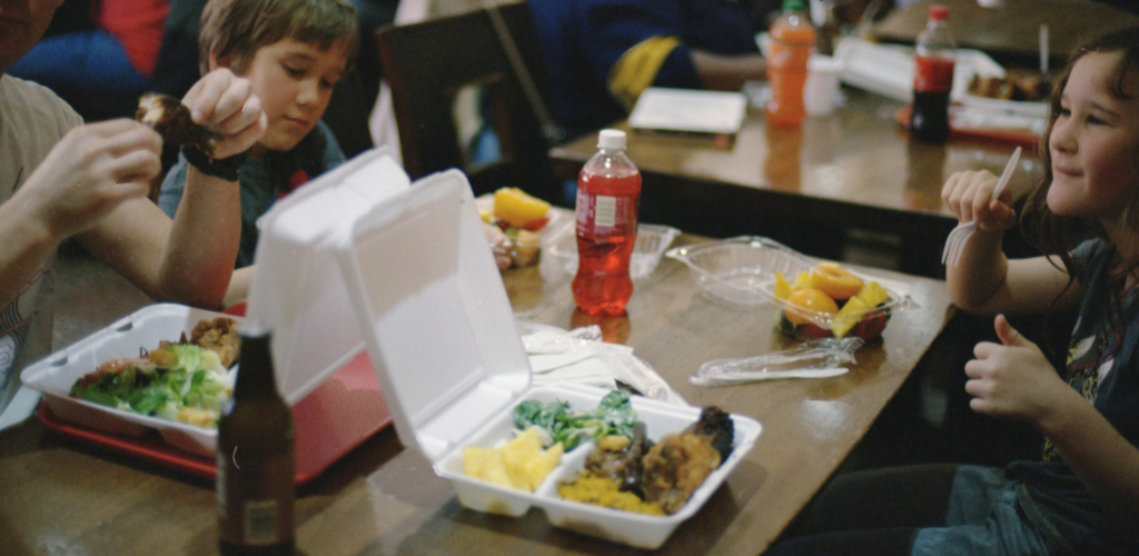 A group of children sit at a wooden table eating takeaway meals from white foam containers. The plates show fried chicken, greens and pineapple; bottled sodas, clear plastic fruit cups and utensils are scattered on the table. One child holds a drumstick while another gives a thumbs-up.