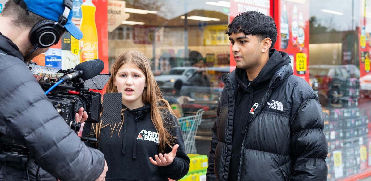 Young activists Maya and Dev are standing in front of a store with cans of coke on display. They are speaking to camera.