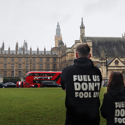 2 young people with their backs to the camera are facing Westminster building. They are wearing black Bite Back hoodies which have white text on the back that says, 'Fuel us, Don't fool us'