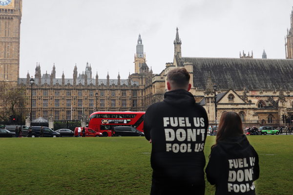 2 young people with their backs to the camera are facing Westminster building. They are wearing black Bite Back hoodies which have white text on the back that says, 'Fuel us, Don't fool us'