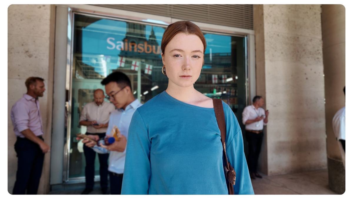 Young woman with reddish hair and a blue top looks directly at the camera while standing outside a Sainsbury’s storefront. Blurred commuters and stone columns fill the background; she has a brown shoulder bag.