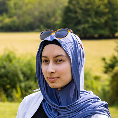 Zainab Headshot Serious:A young woman with a blue headscarf wrapped loosely and glasses perched on her head, is looking into the distance with a serious expression.