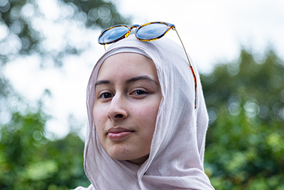 A young woman wearing a white headscarf with sunglasses perched on top is looking into the camera.