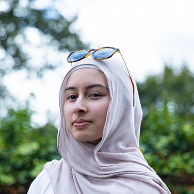 A young woman wearing a white headscarf with sunglasses perched on top is looking into the camera.