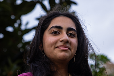 A young South Asian woman with long curly hair is looking into the camera with a serious expression.