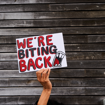 A hand holding a sign that says 'We're biting back' in large, bold, black and red writing that has been painted on. The background is wooden slats on the wall of a building.
