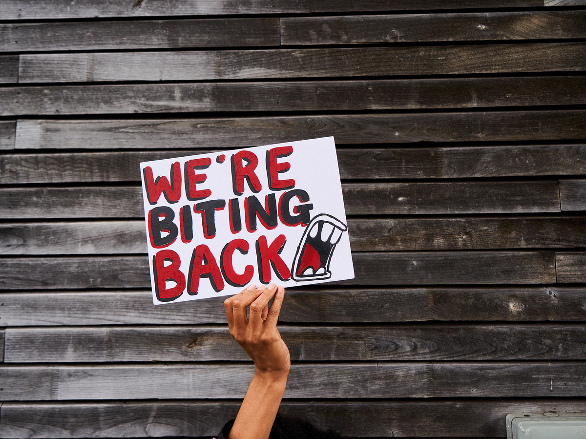 A hand holding a sign that says 'We're biting back' in large, bold, black and red writing that has been painted on. The background is wooden slats on the wall of a building.